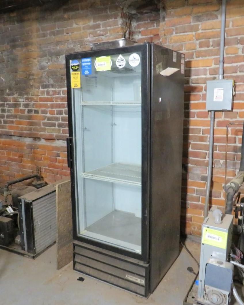 Tall, black commercial refrigerator with glass door inside a brick-walled room.