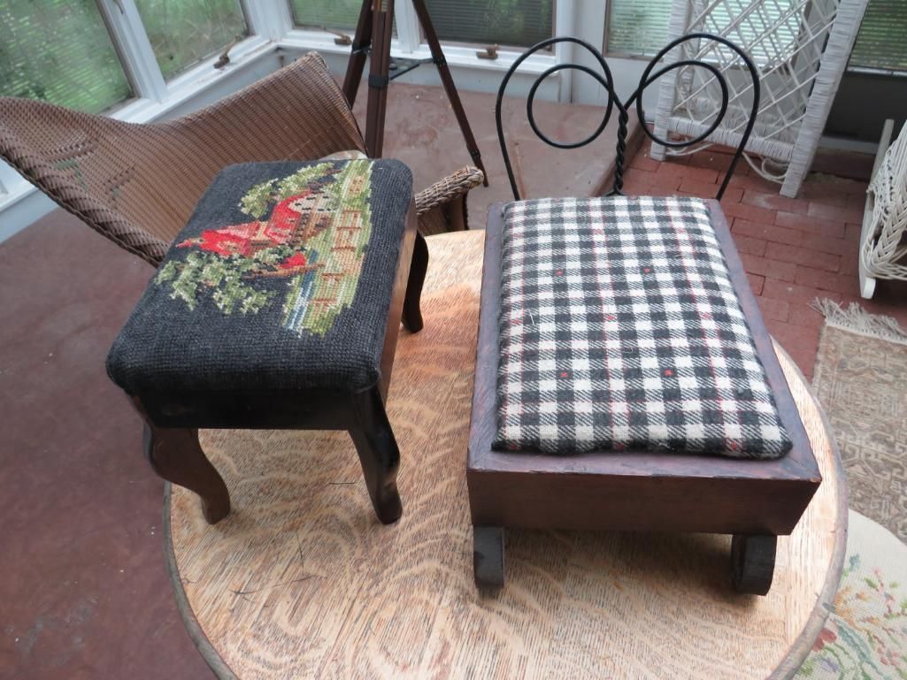 Two upholstered footstools on a round table; one with a plaid top and one with a needlepoint scene.