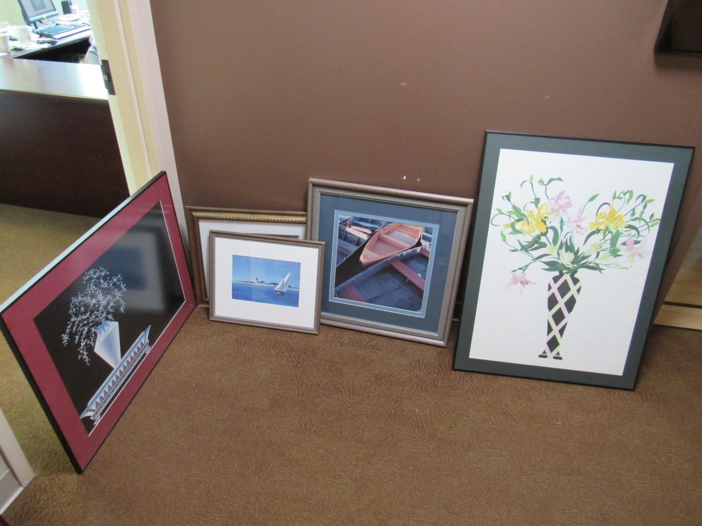 Five framed art pieces leaning against a brown wall and beige carpet.