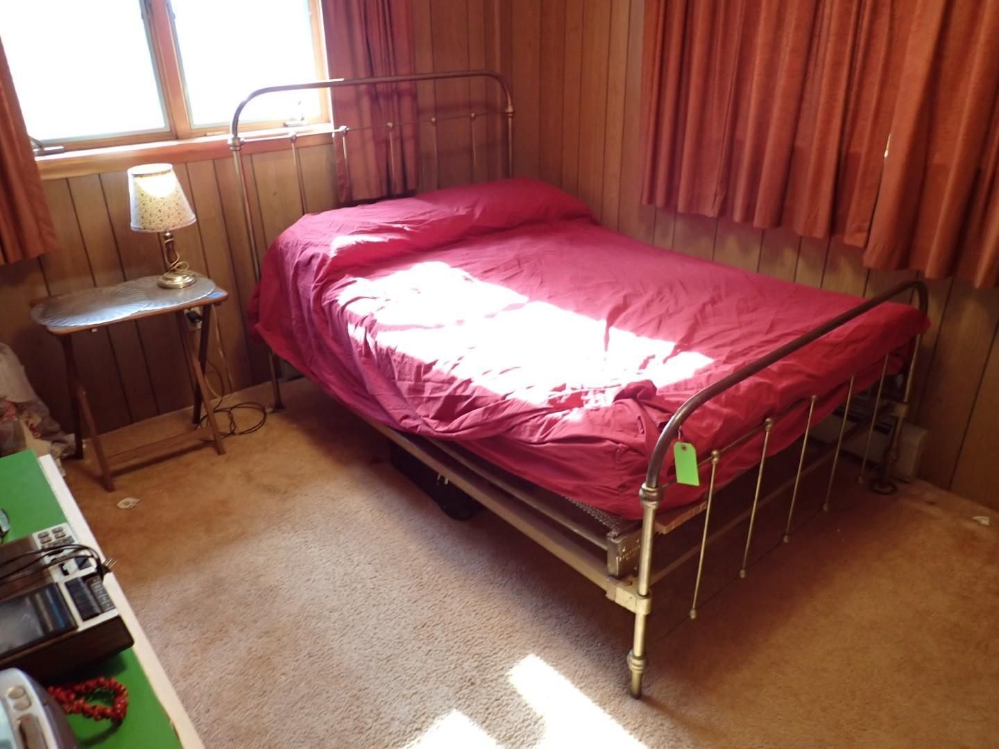 A metal bed frame with a red comforter sits in a room with wood-paneled walls and a small side table.