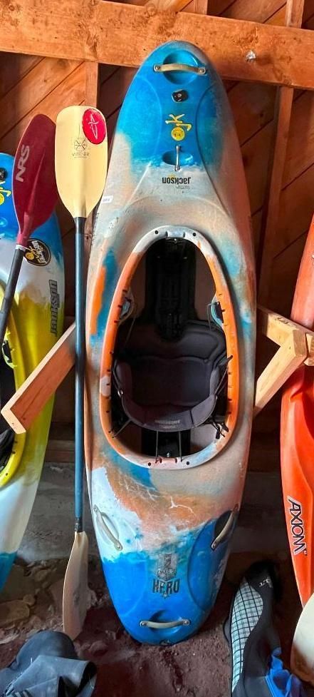 A kayak, with paddle, hanging in a wooden storage area. Blue, tan, and orange color scheme.