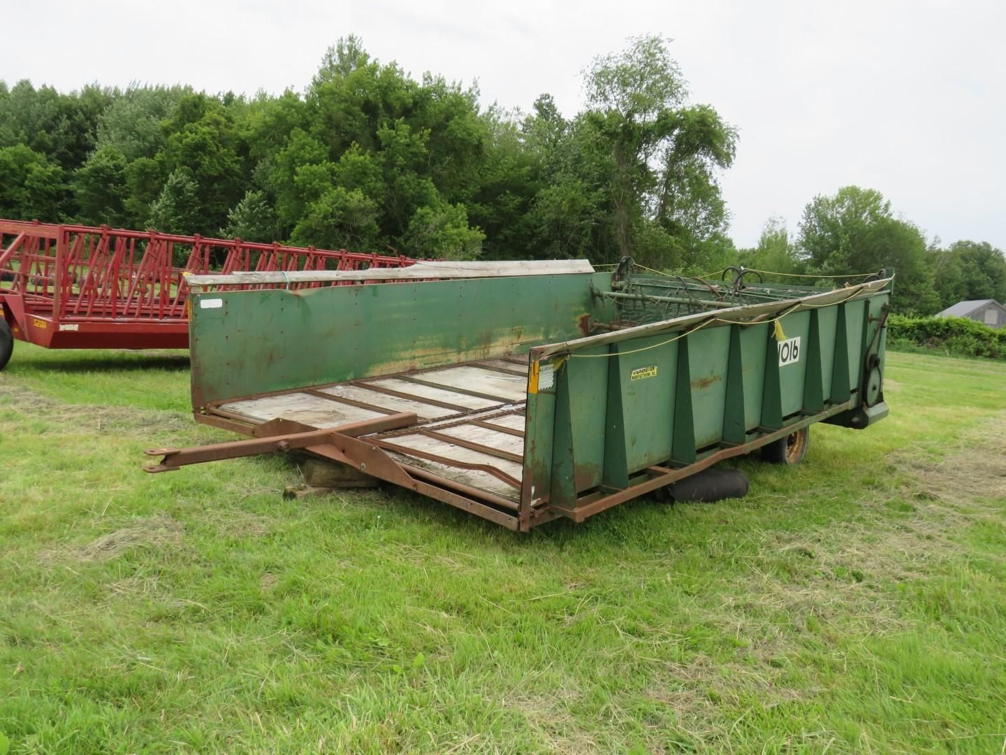 Green farm trailer on a grassy field with a red hay wagon in the background.