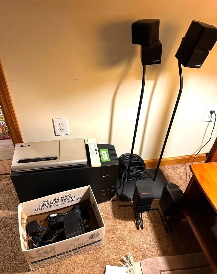 Bose stereo speakers with stands, a control console, subwoofer, and accessories in a room with tan walls.