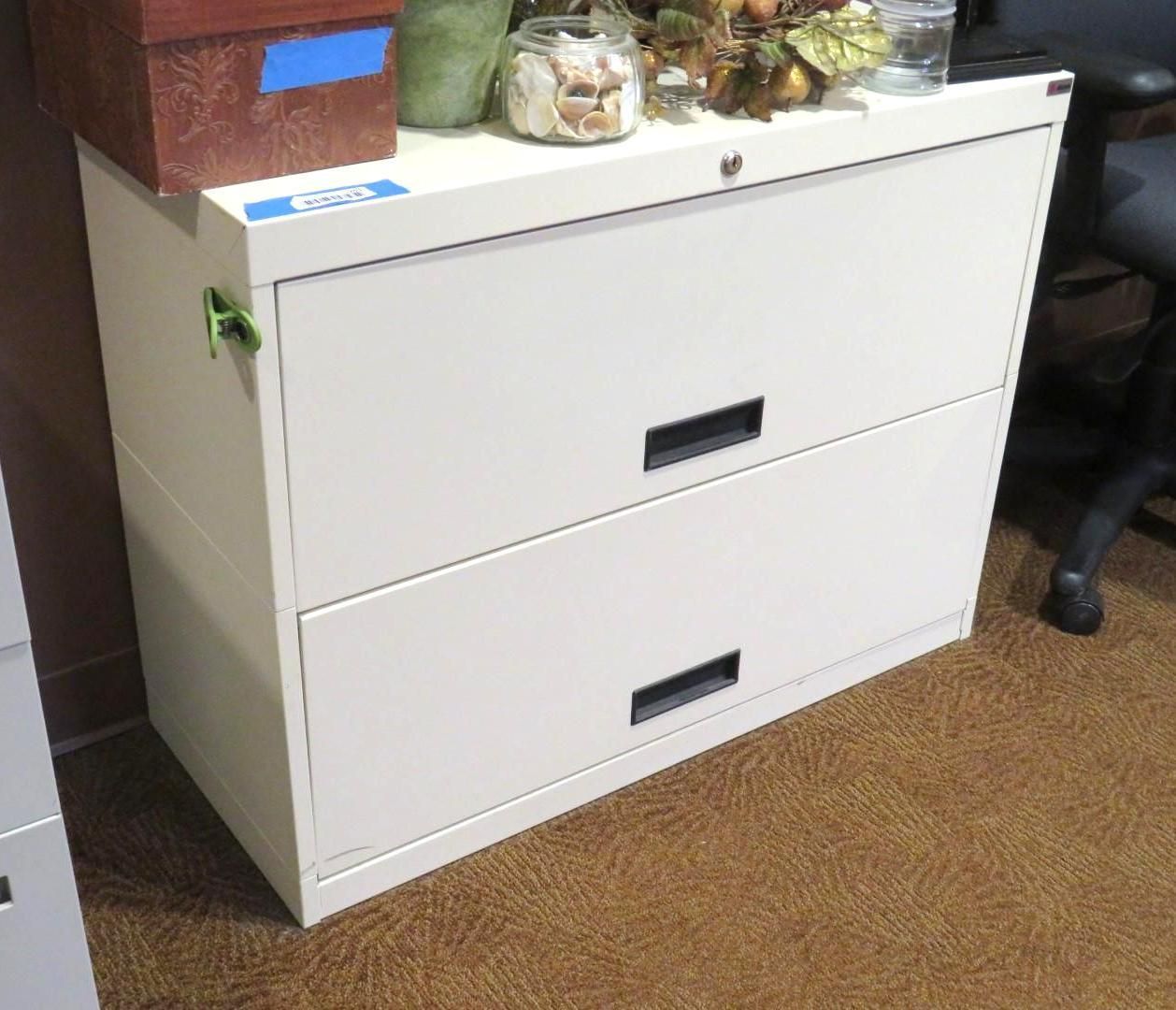Two-drawer cream-colored metal filing cabinet. Top drawer locked, black handles. On brown carpet, items on top.