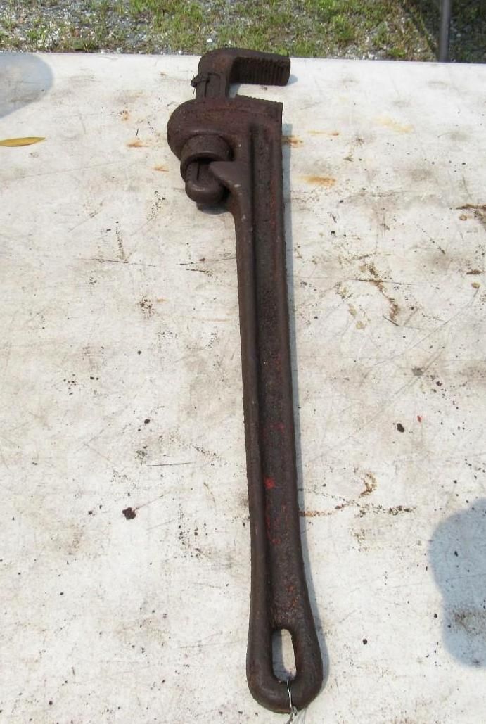Pipe wrench on a light-colored surface. The wrench is dark brown and in focus, with a loop at the handle's end.