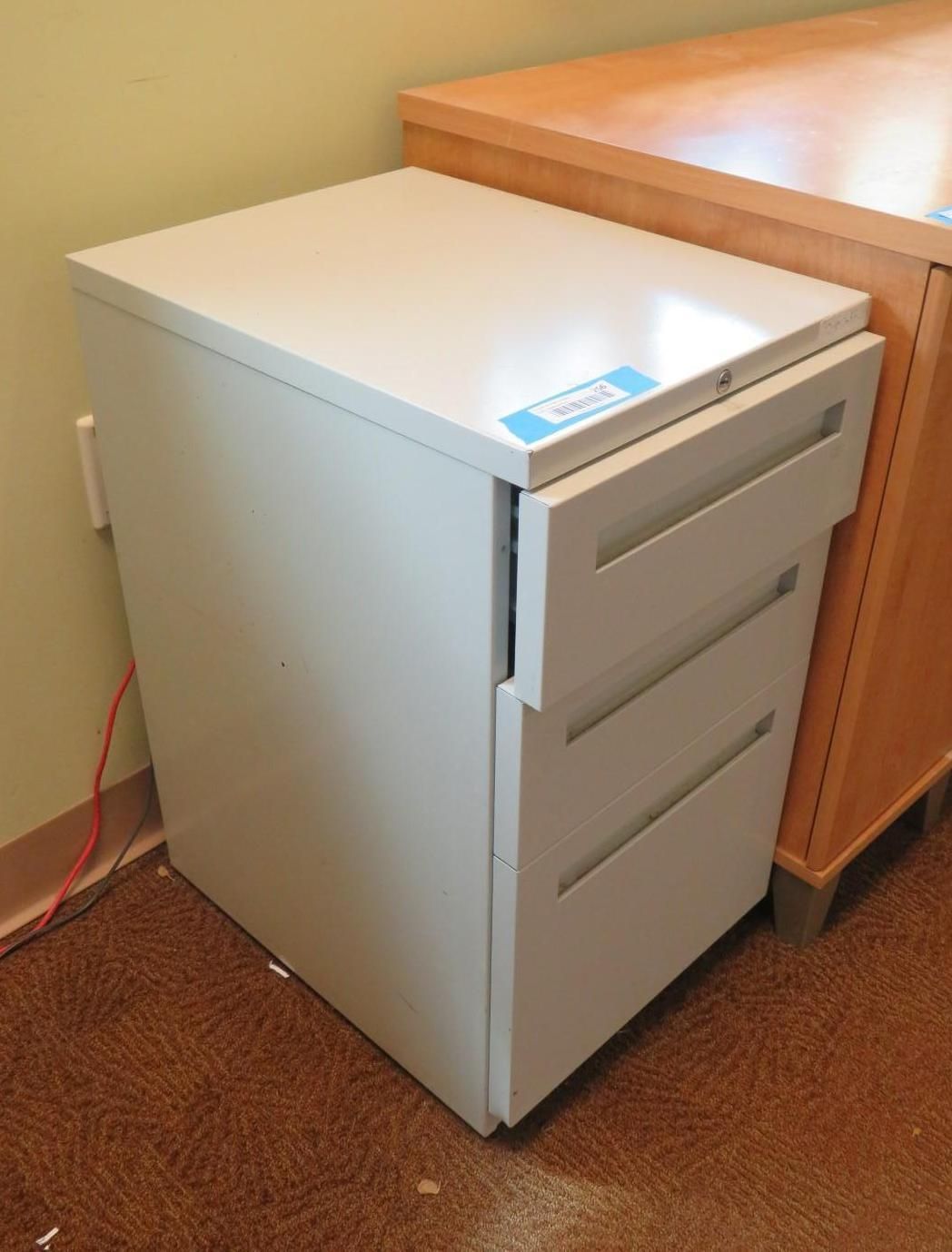 White three-drawer filing cabinet beside a light brown desk; red cable on wall, brown carpet.