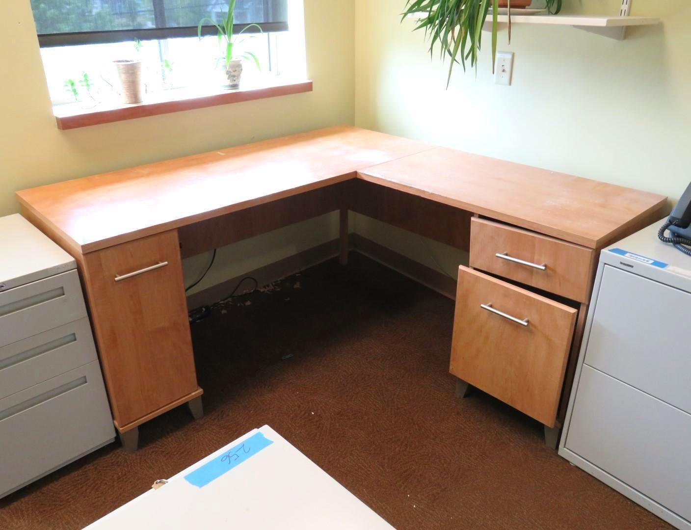 L-shaped wooden desk in office setting, two filing cabinets on either side.