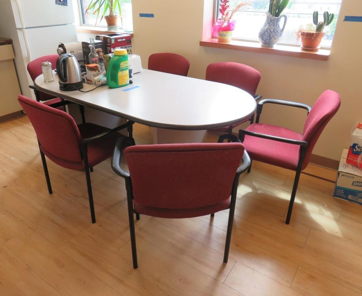 Oval conference table surrounded by six red chairs, near a window with plants.