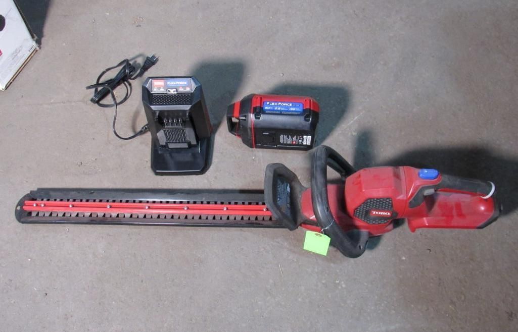 Red and black cordless hedge trimmer with charger and battery on a gray surface.