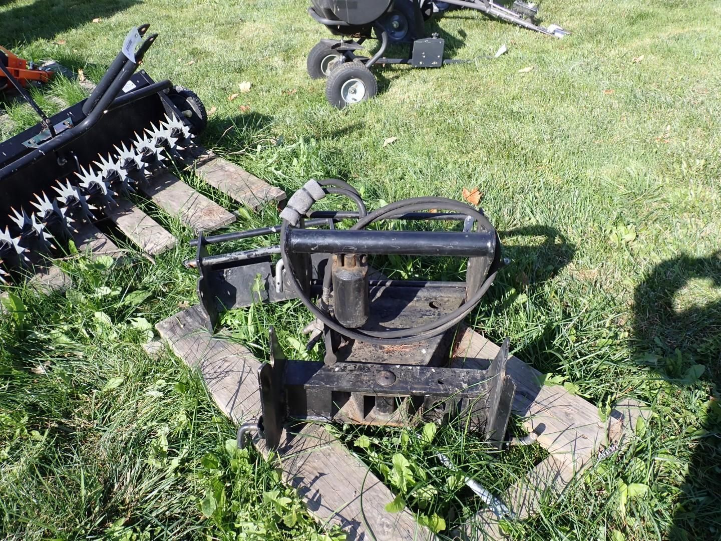 Black metal farm implement with tines and hose reel resting on grass and stones outdoors