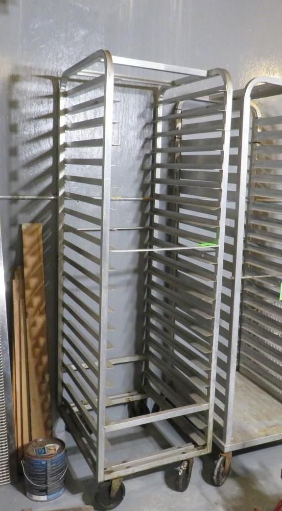 Metal bakery rack on wheels. Multiple shelves for trays. Standing near a wall.