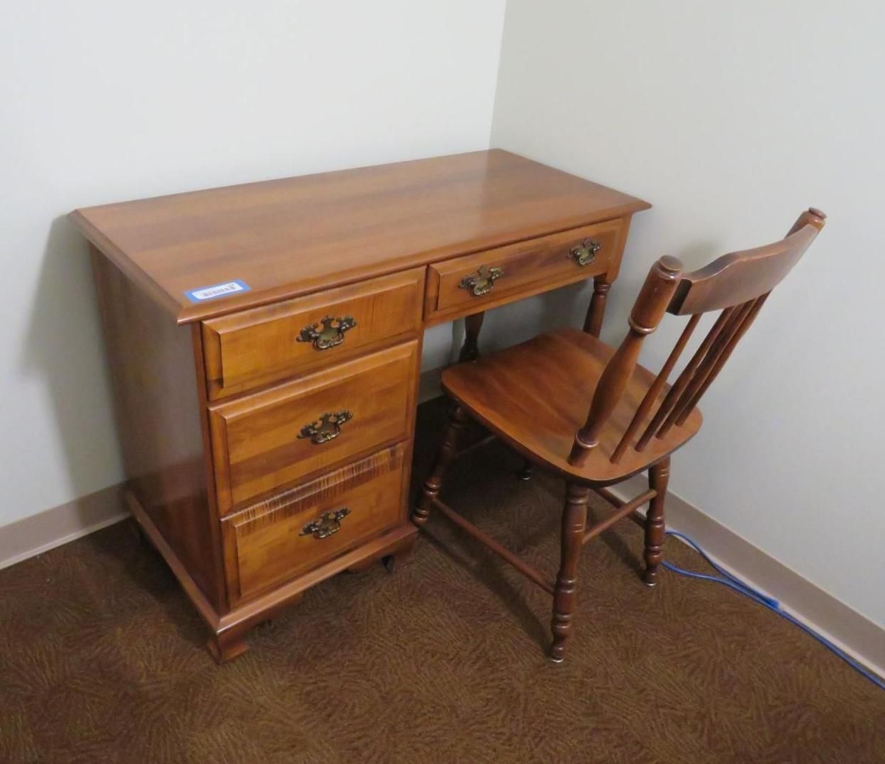 Wooden desk and chair in a corner of a room, brown and tan hues, side drawers, and a wooden back chair.