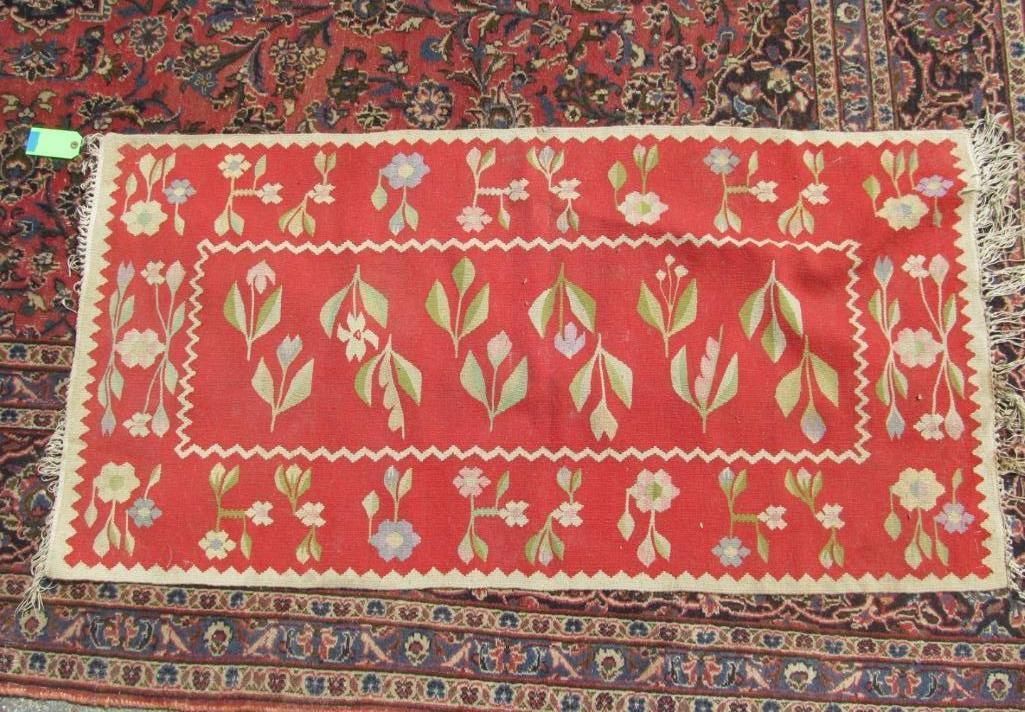 Red rug with floral and leaf designs, fringed edges, atop another rug with a dark pattern.