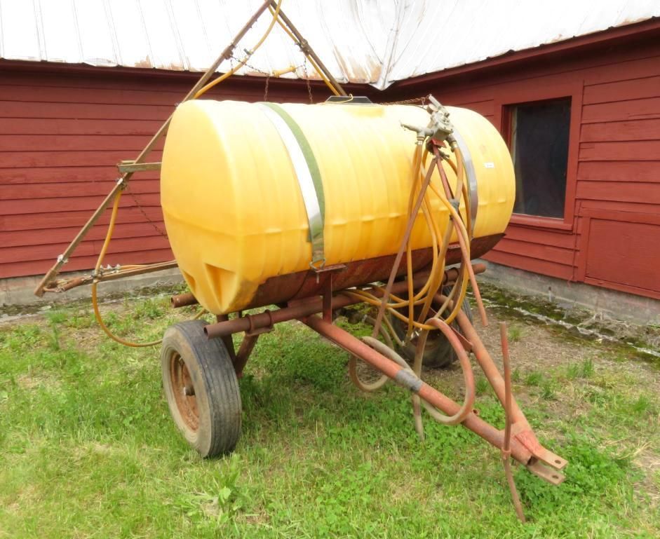 Yellow agricultural sprayer tank on a red metal trailer, parked on grass near a red building.