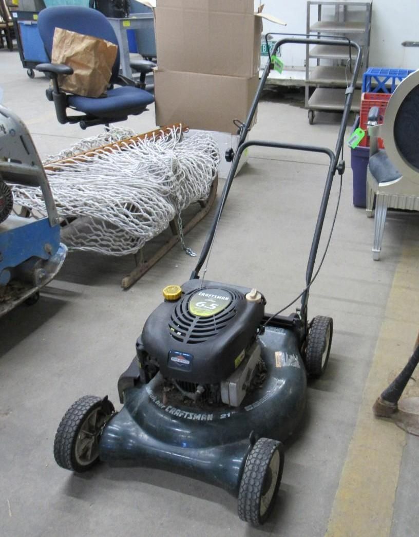 A lawnmower with a black engine and handle, gray body, and black tires, indoors.