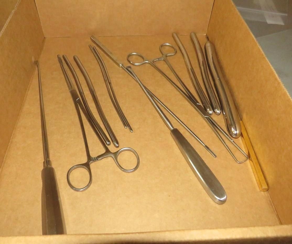 Surgical instruments in a brown cardboard box, including forceps, scissors, and a needle holder.