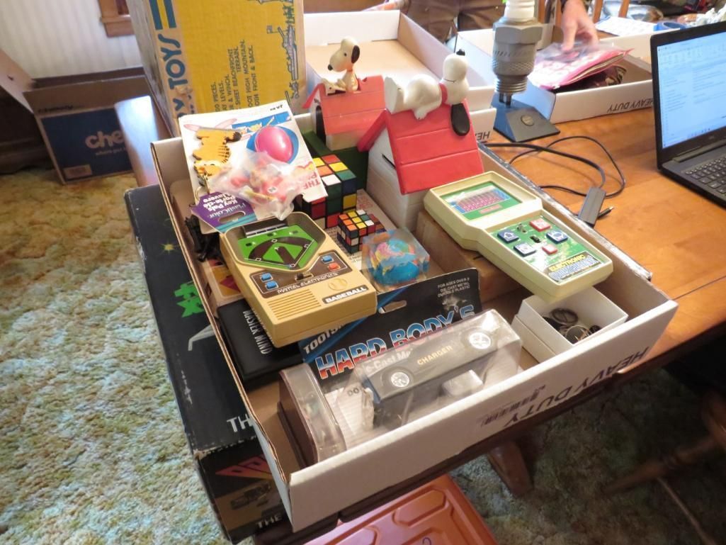 Cardboard box on a table filled with vintage toys: handheld game, Rubik's cube, dog figurines, and packaging.