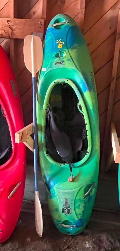Green and blue kayak with a paddle hanging in a wooden structure next to a red kayak.