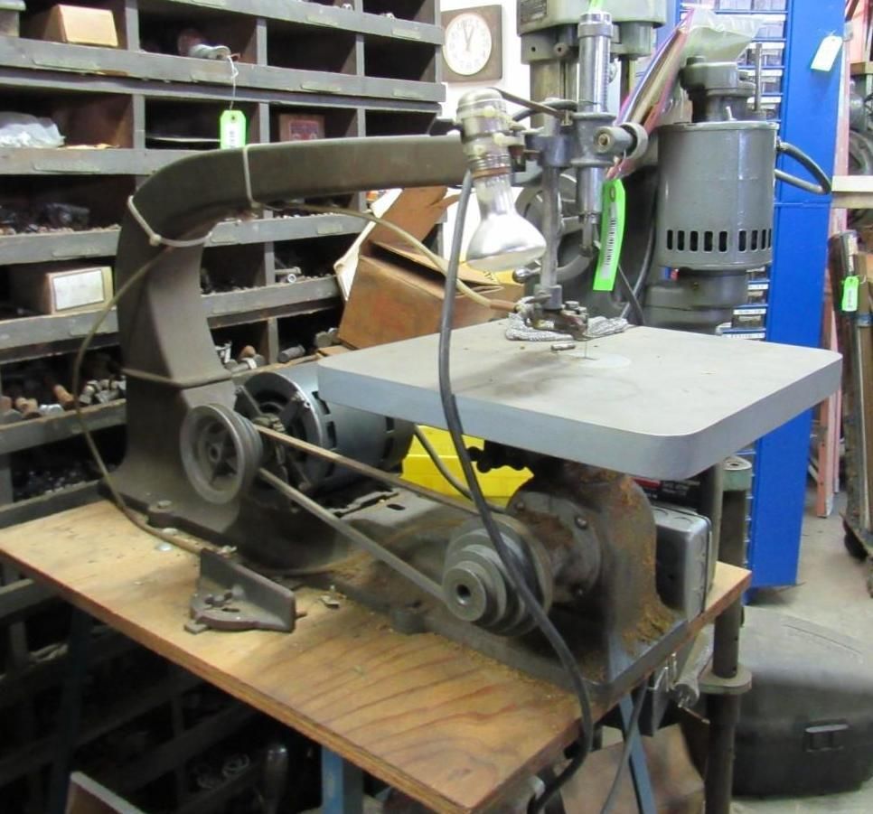 Gray scroll saw machine on a wooden table, with pulleys, belts, and a light.