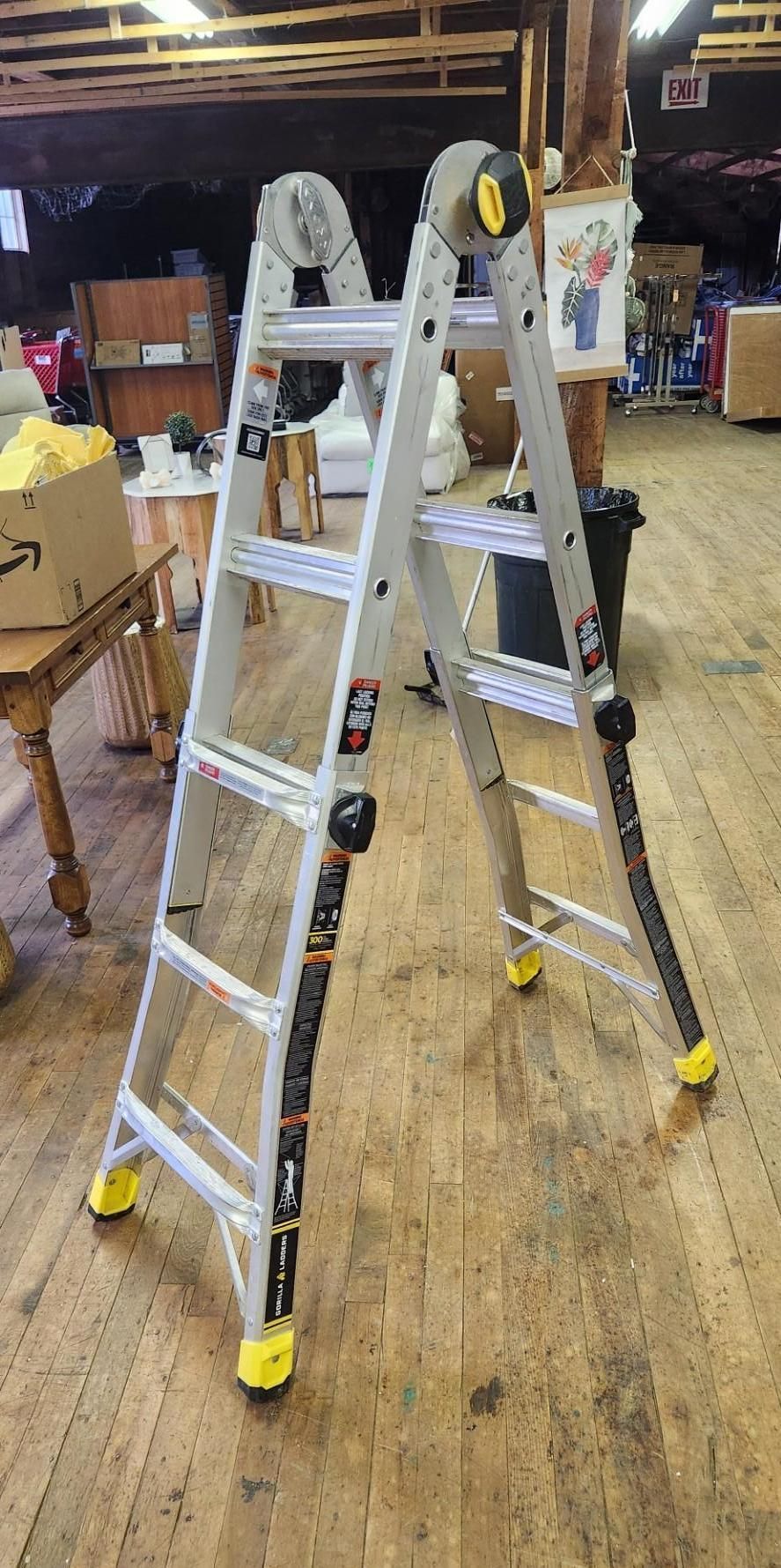 A metal multi-position ladder set up on a wooden floor in a room. The ladder is silver with yellow feet.