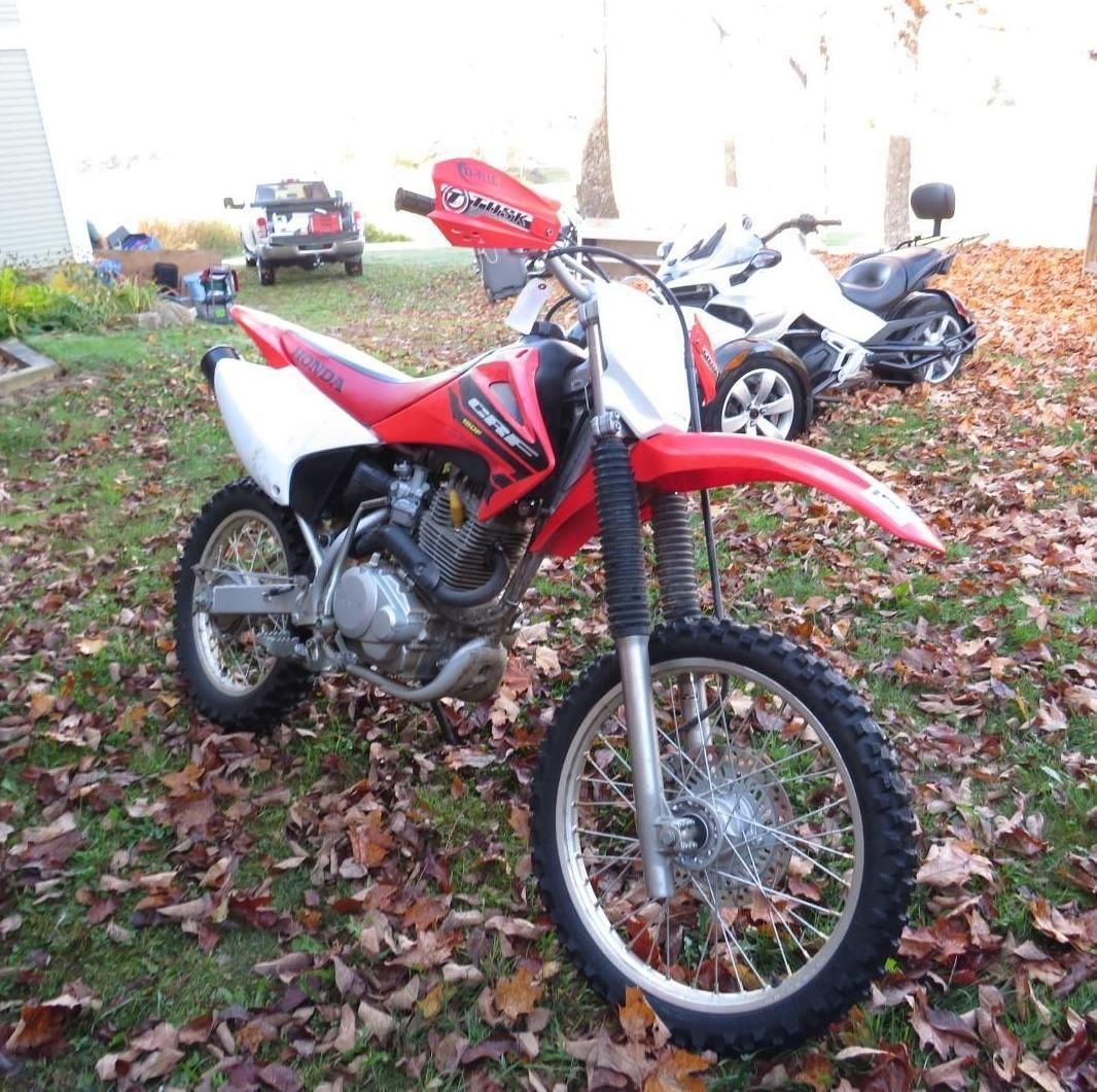 Red and white Honda dirt bike on grass, surrounded by fallen leaves, with other vehicles in the background.