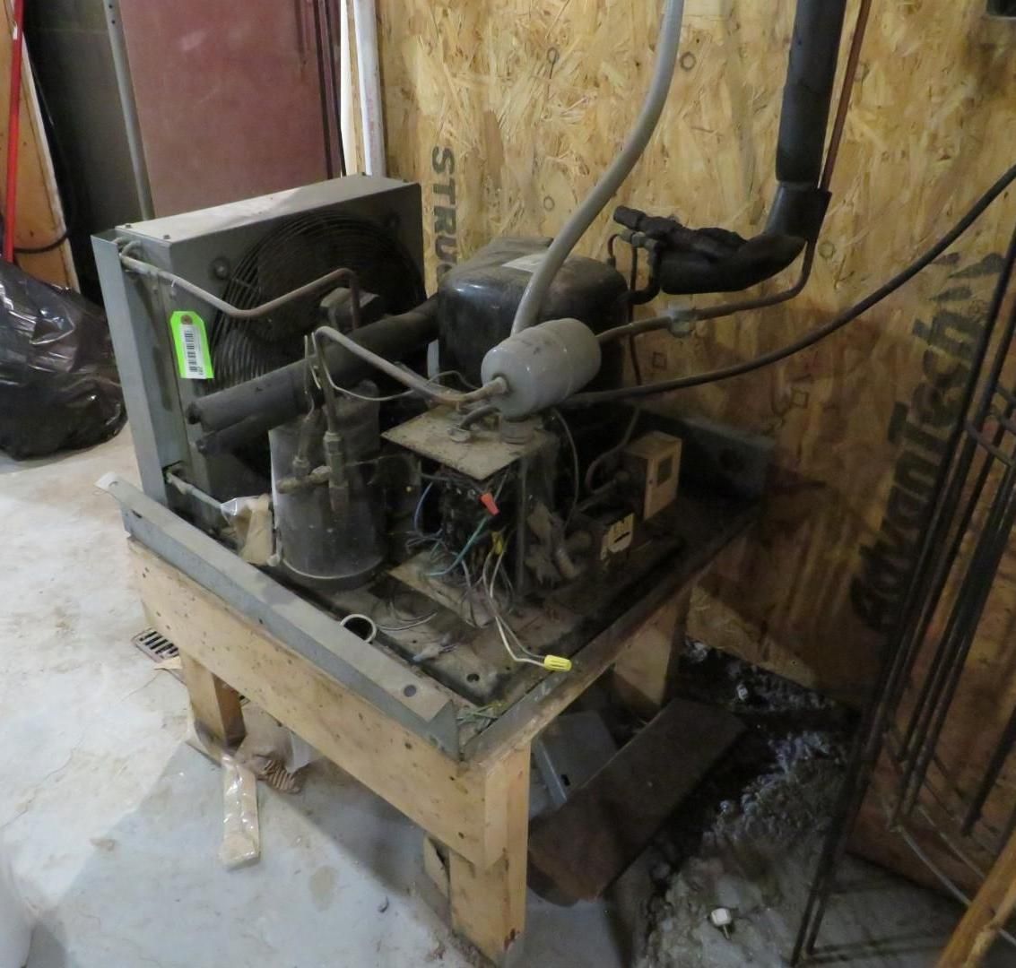 A refrigeration unit sits on a wooden stand in a room with plywood walls.