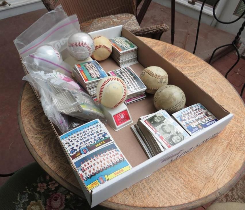 A cardboard box on a table contains baseballs and stacks of baseball cards.