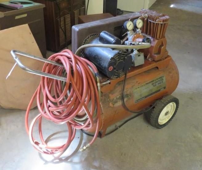 Orange air compressor with gauges, hose, and wheels; indoors.