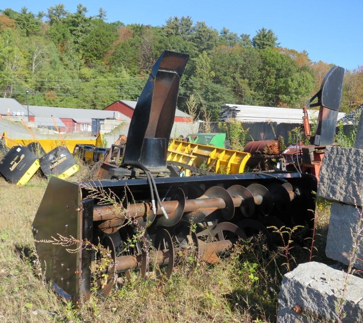 A snowblower sits with industrial buildings and trees in the background.