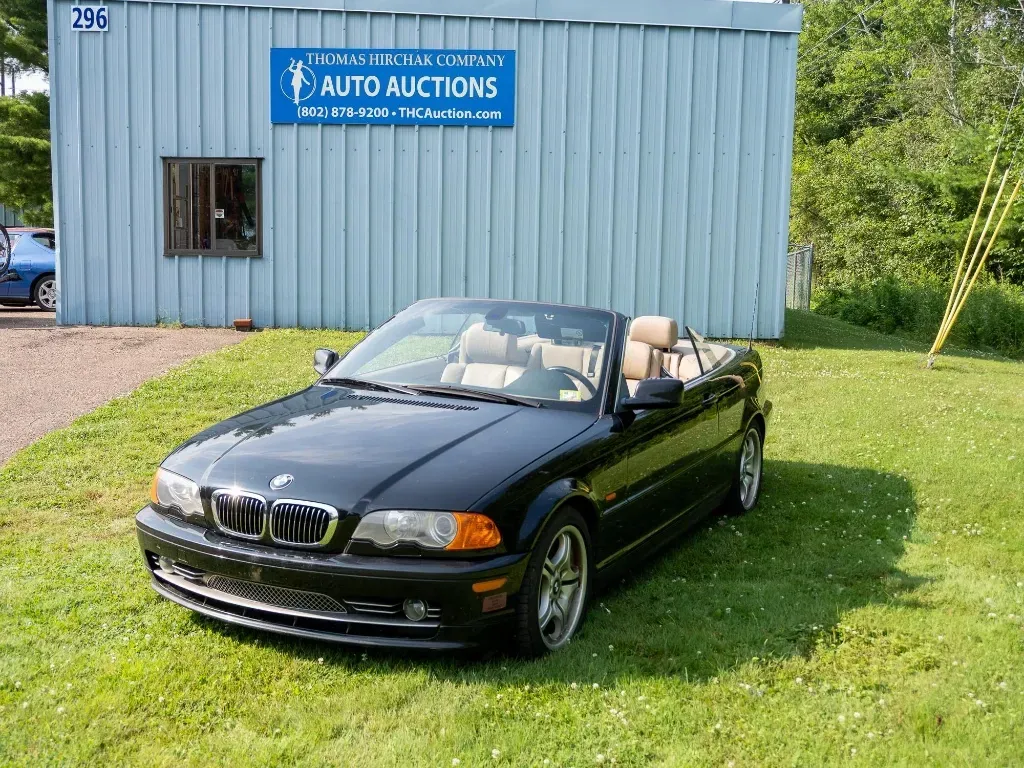 Black BMW convertible in front of a blue auto auction building, on green grass.