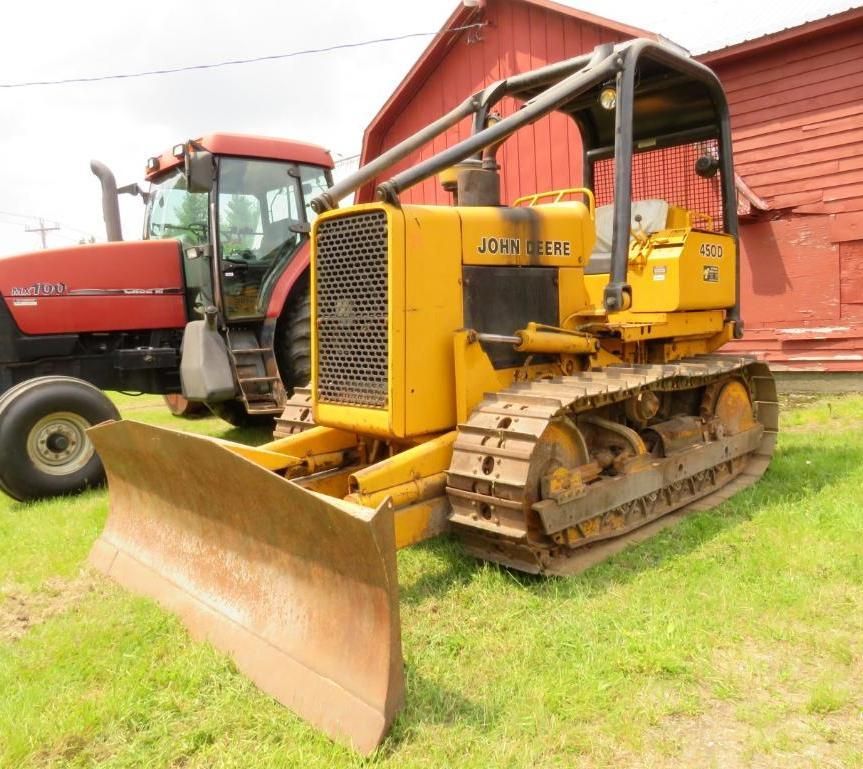 Yellow John Deere bulldozer with blade on grassy field, red tractor in background.