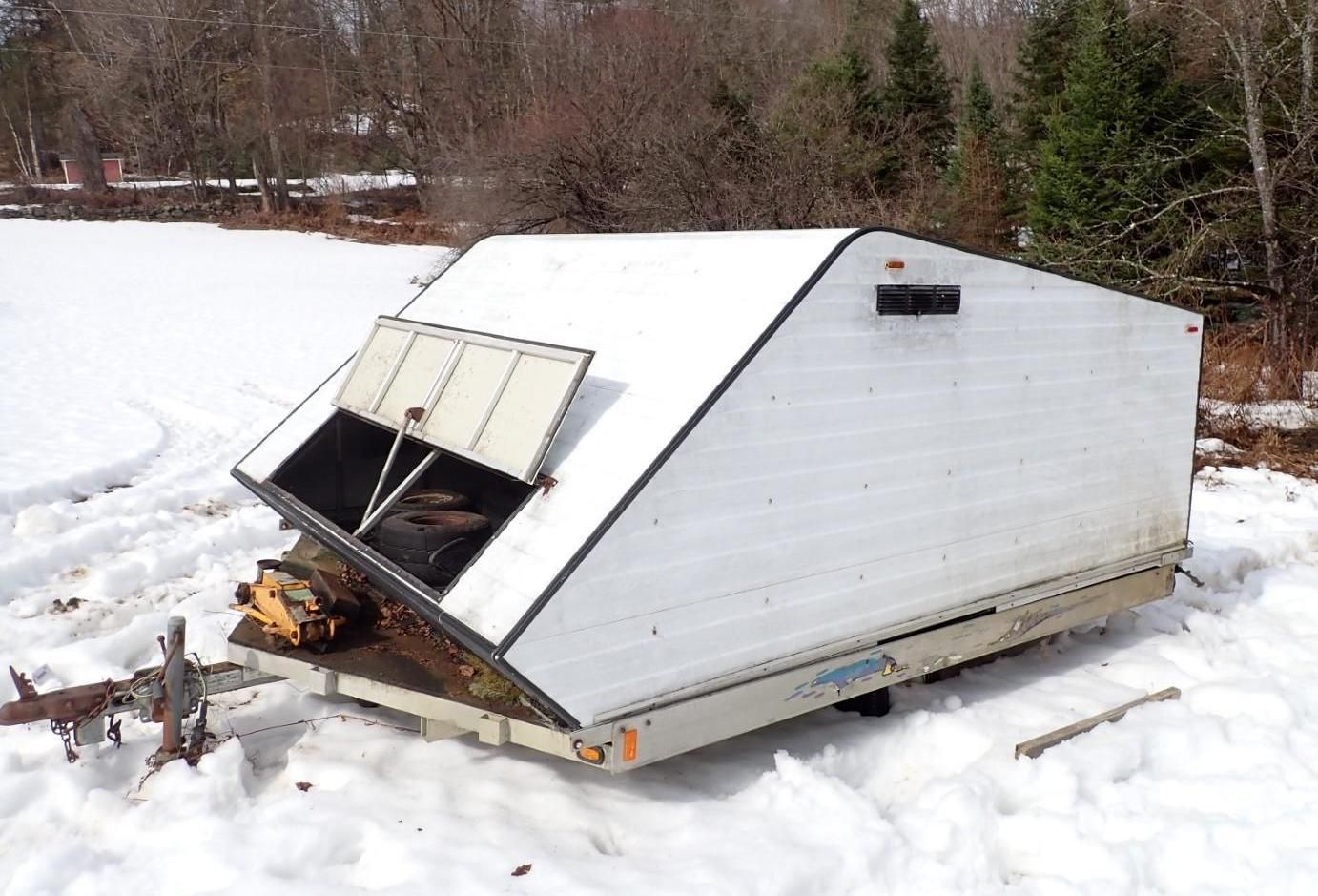 A white snowmobile trailer with a raised storage compartment hatch, parked on a snowy, wooded landscape.