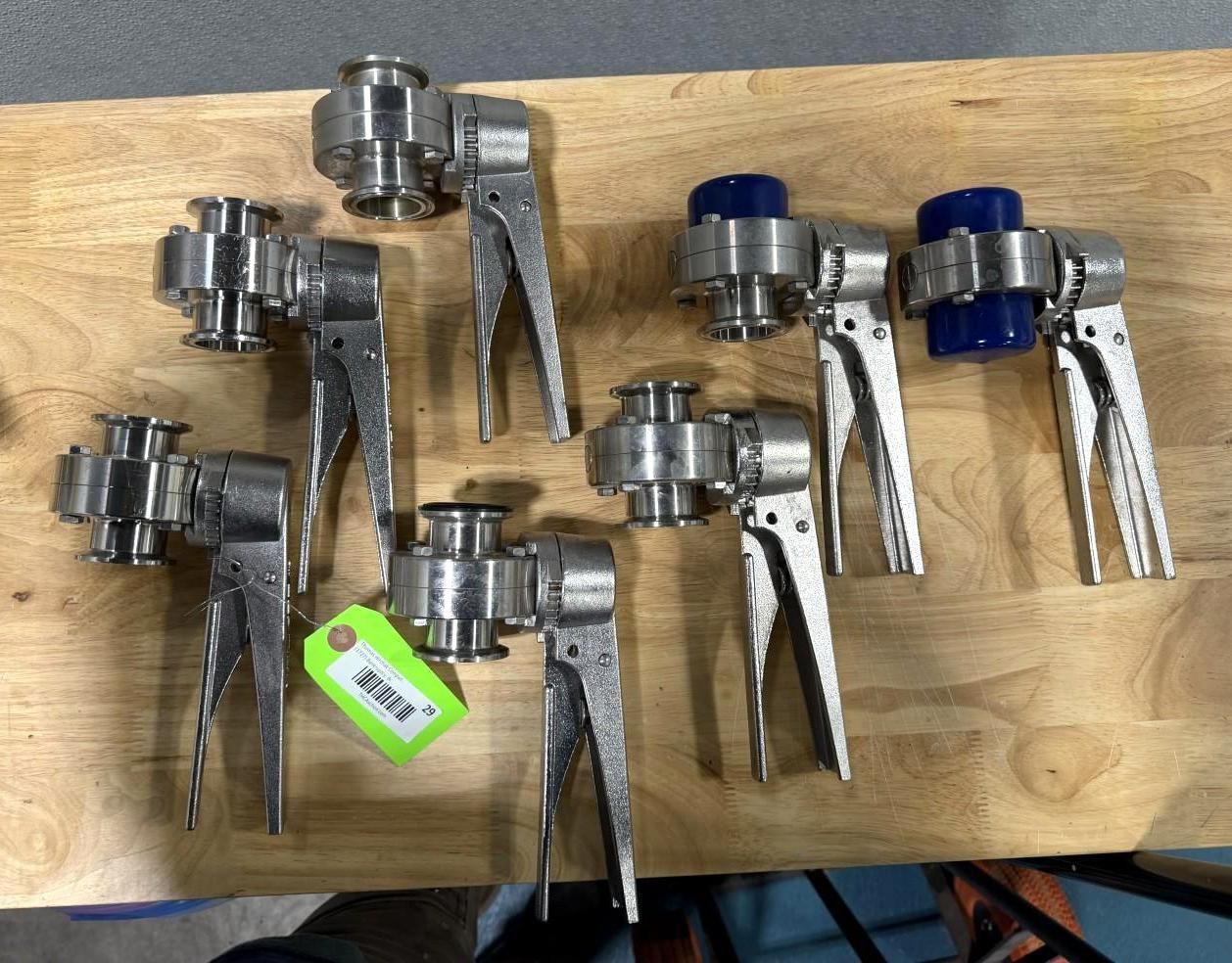 Seven stainless steel butterfly valves with lever handles on a wooden surface. Two have blue caps.