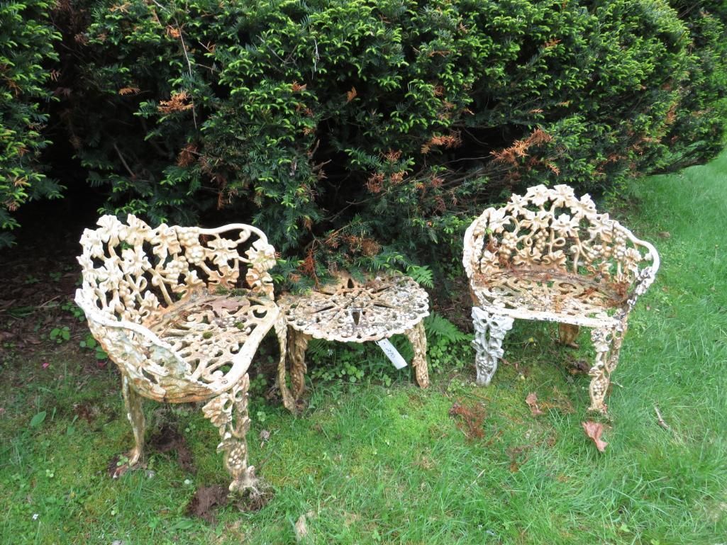 Three ornate, white, metal patio chairs and a table sit on grass in front of a green bush.