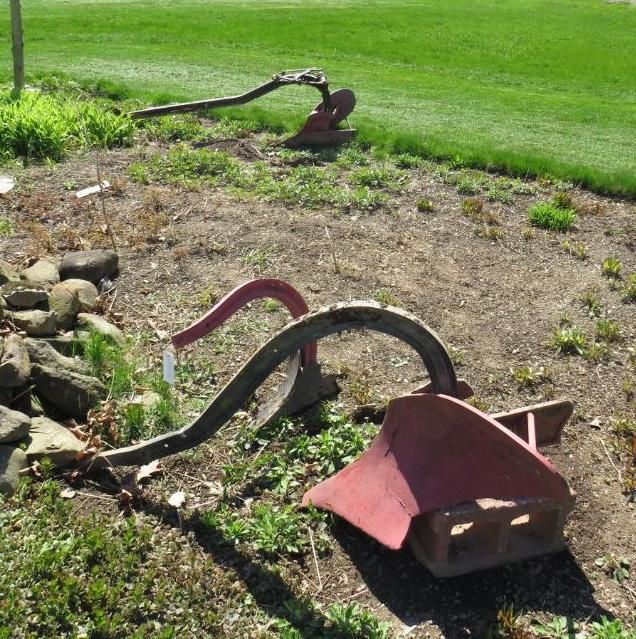 Two antique red plows rest in a garden bed near green grass.