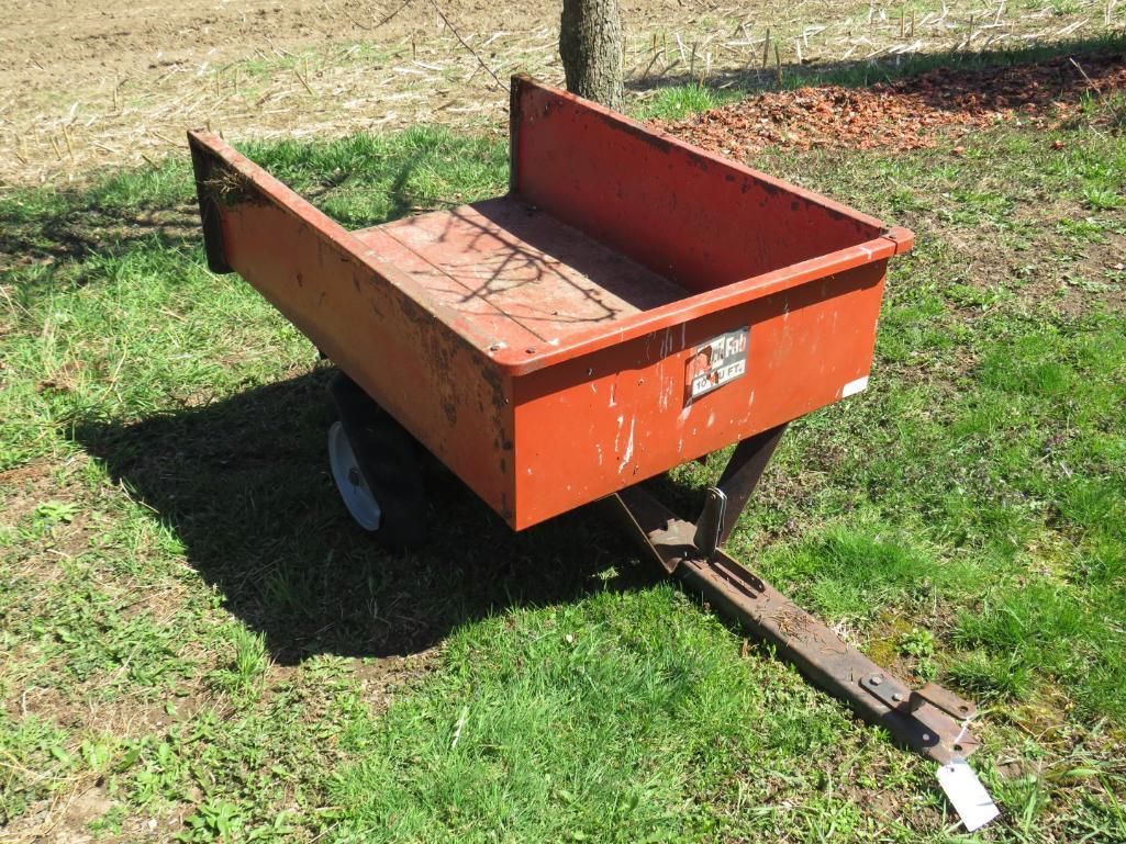 Orange metal utility cart on wheels in a grassy field, attached to a tow bar.