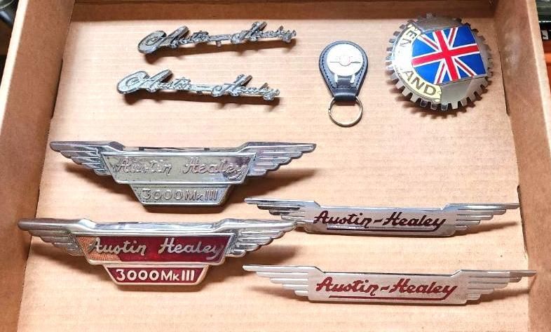 A wooden box displays vintage Austin-Healey car emblems: multiple logos, a keychain, and a circular sign with a Union Jack.