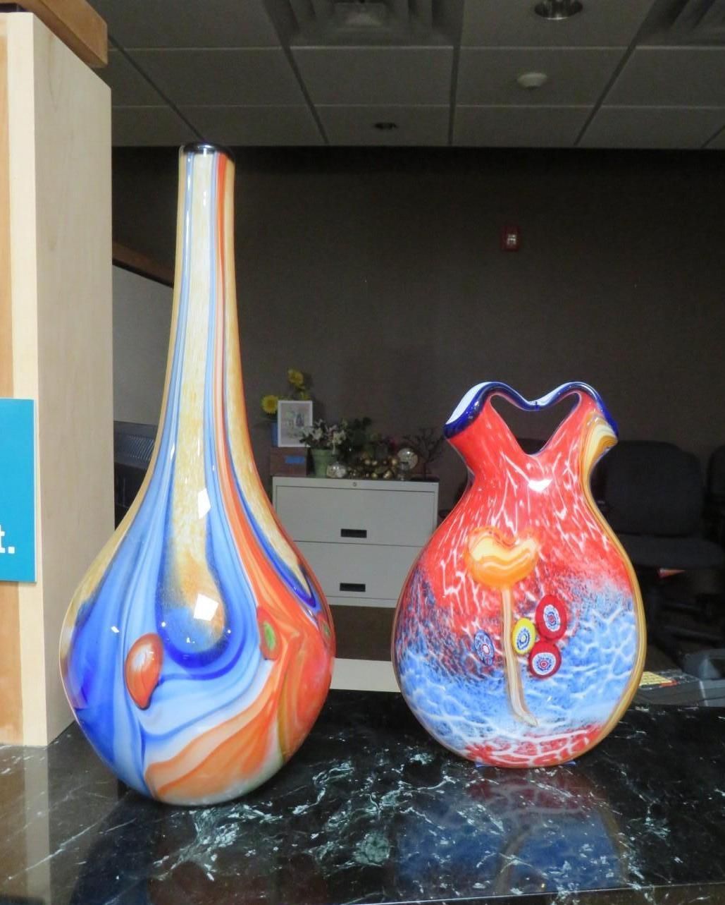 Two colorful glass vases on a black surface; the left is tall and pear-shaped, the right is short with a ruffled rim.