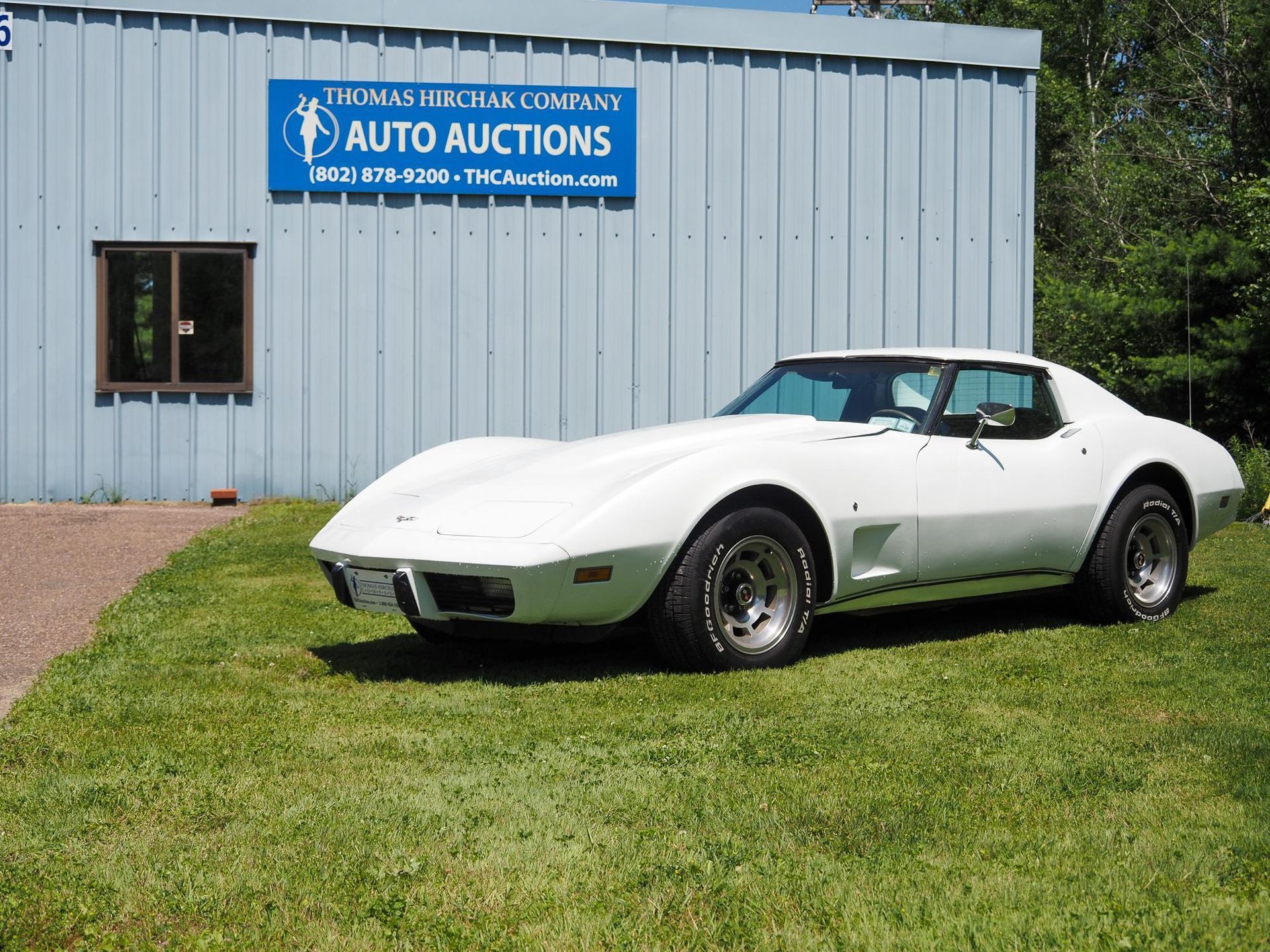 White Corvette sports car parked on grass in front of a blue-gray building with 