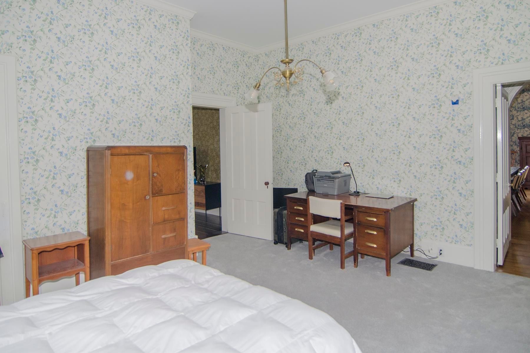 Bedroom with floral wallpaper, wooden furniture: wardrobe, desk, nightstand. A bed is in foreground.