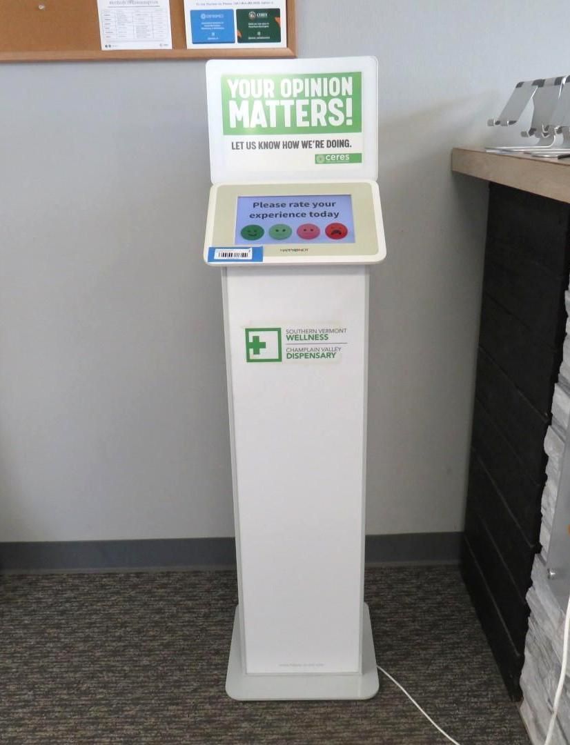 White customer feedback kiosk with a screen and buttons that invites users to share their opinions.