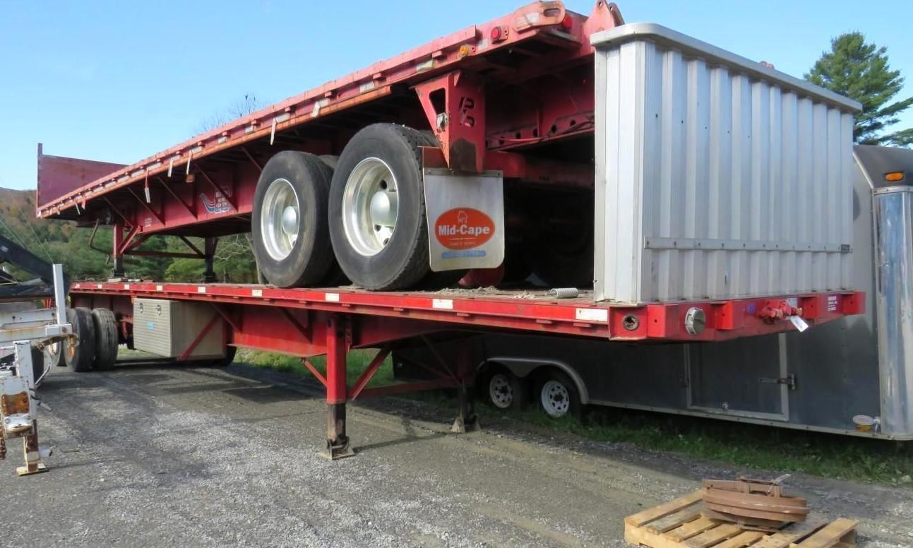 Red flatbed trailer with silver container, parked outdoors on gravel.