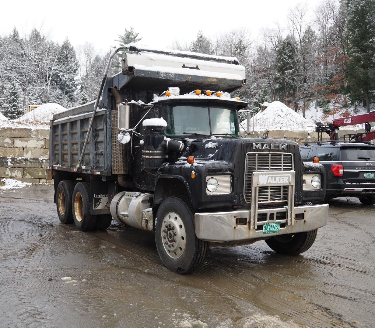 Black Mack dump truck parked on a snowy lot. Gray cab and bed. Winter setting.