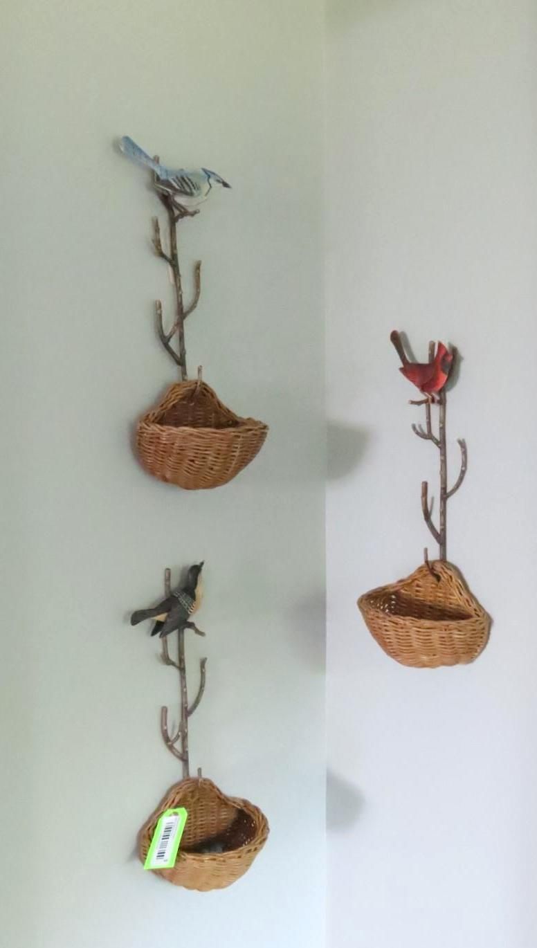 Three wall-mounted bird feeders, each with a metal branch and a bird perched above a woven basket.