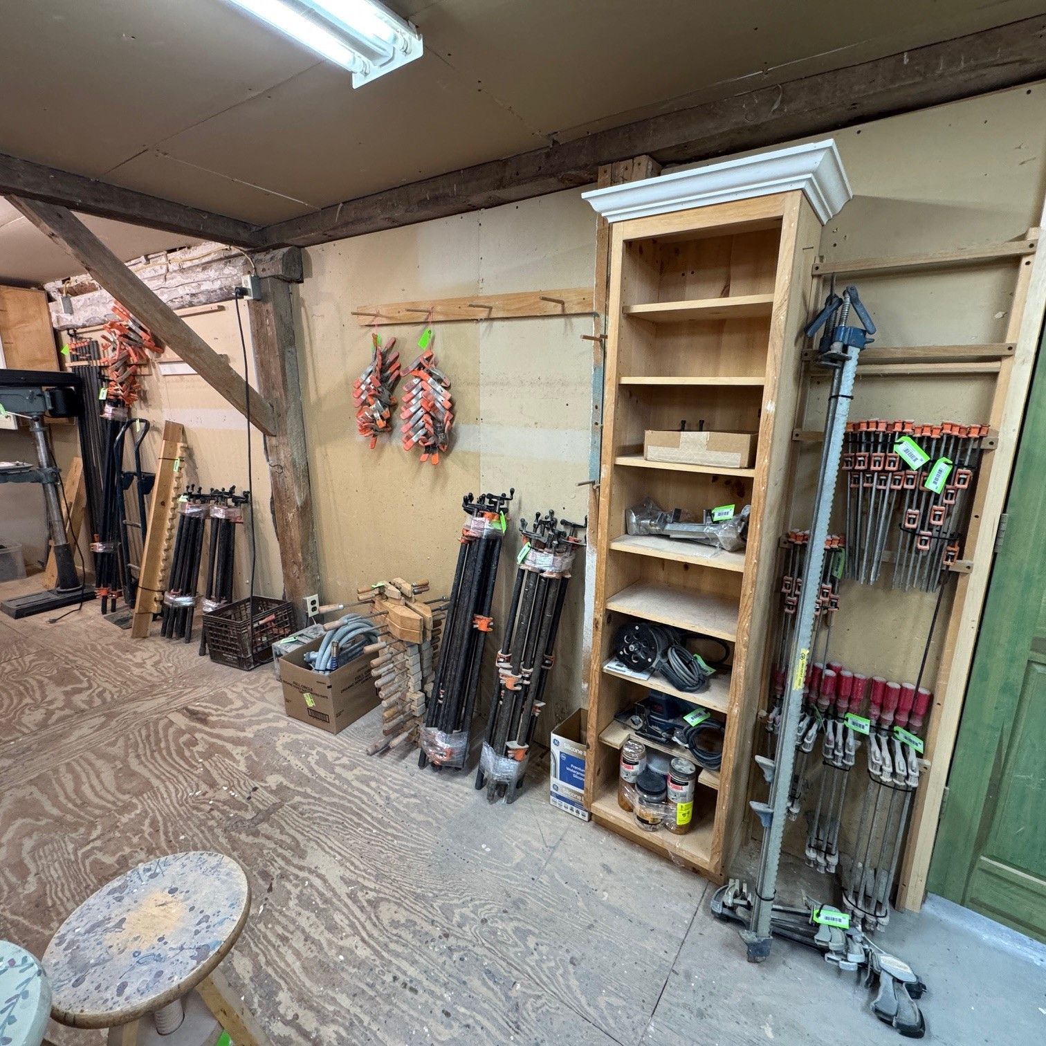 Workshop/storage room with tools, shelves, and rakes against beige walls and a concrete floor.