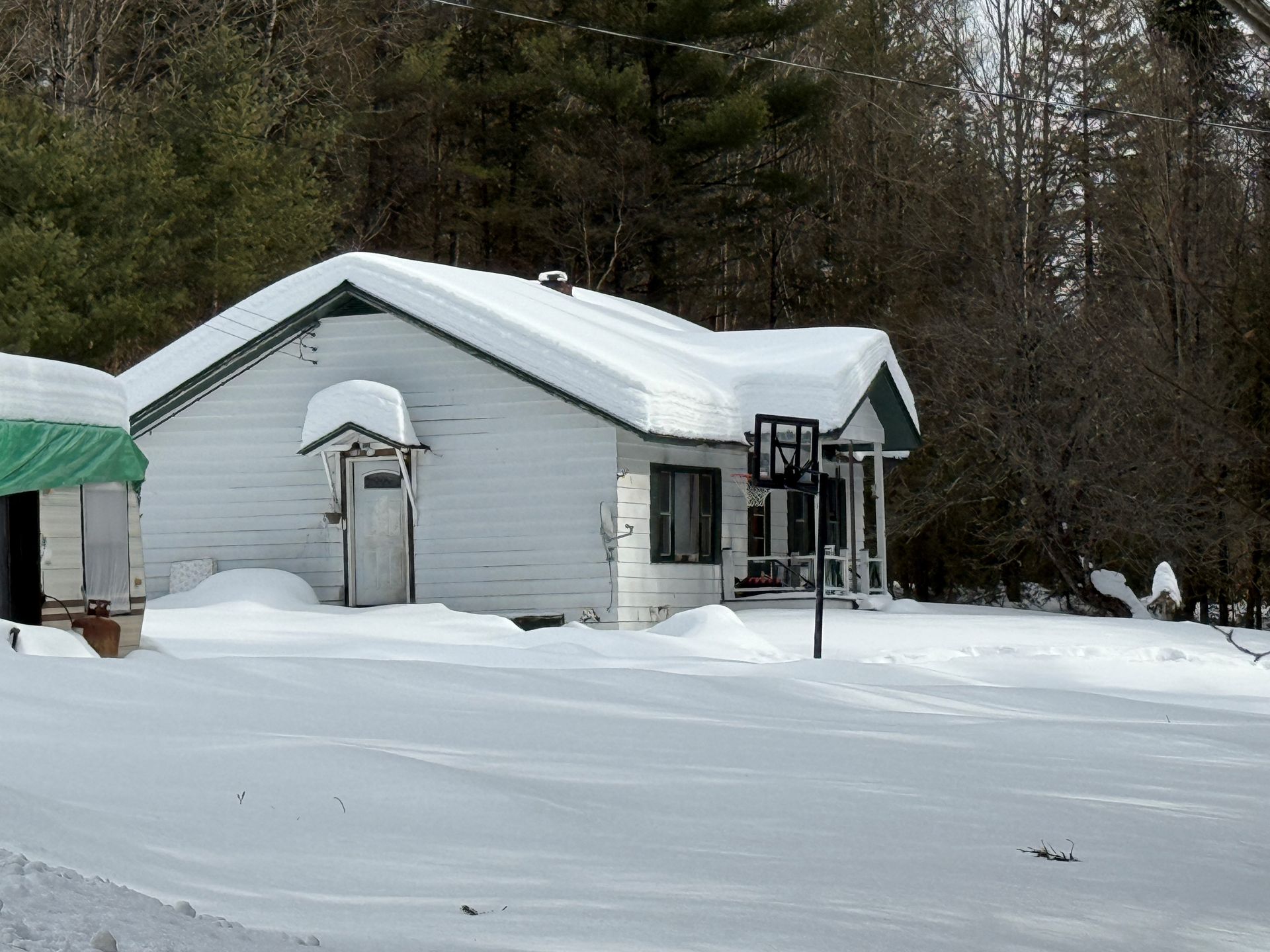 A snow-covered, single-story white house sits in a wooded area with a basketball hoop on the lawn.