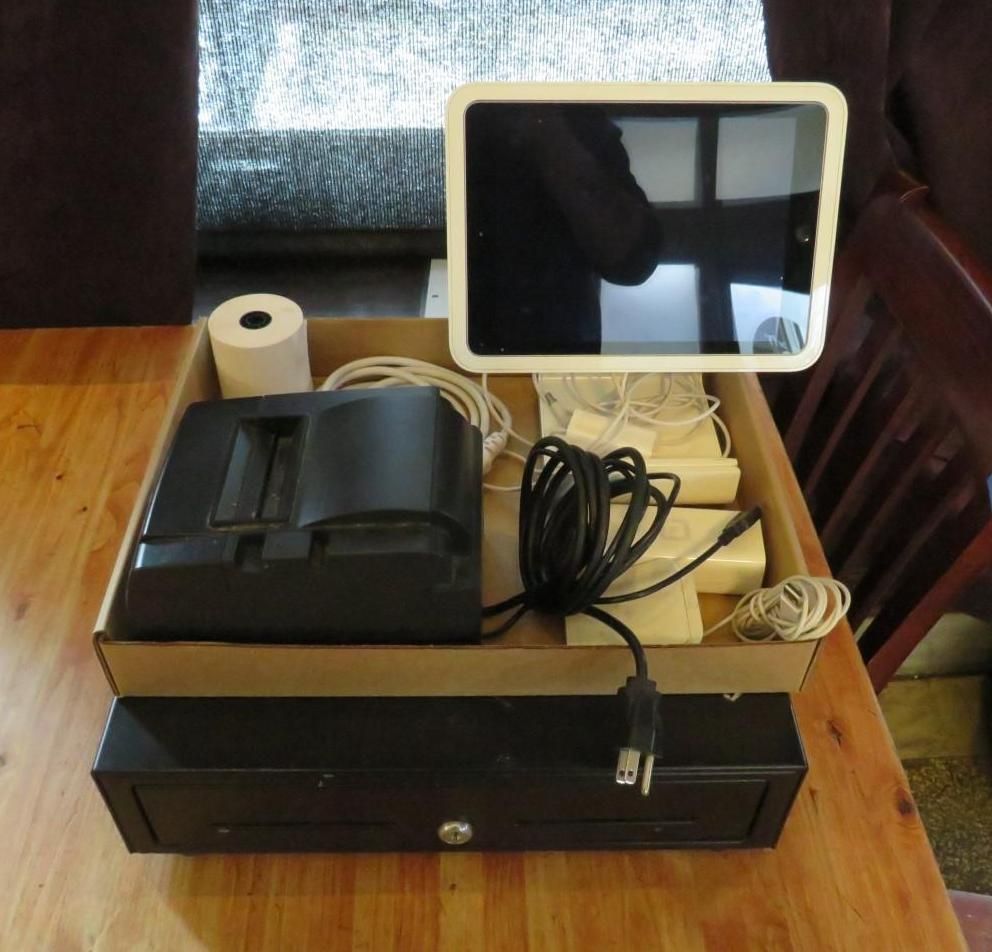 A point-of-sale system on a wooden table includes a tablet, cash register, and receipt printer in a cardboard box.