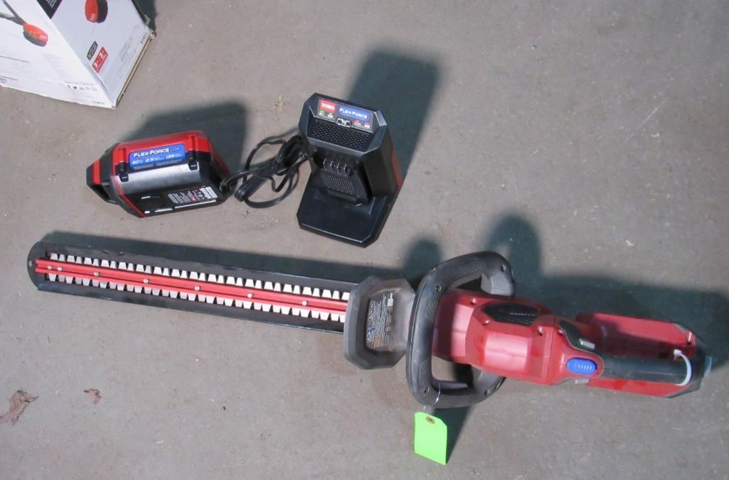 Red and black cordless hedge trimmer with battery and charger, sitting on a floor.