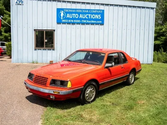 Bright orange Ford Thunderbird parked on grass in front of an auto auction building.