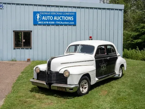 Vintage police car, white and black, parked on grass in front of an auto auction building.